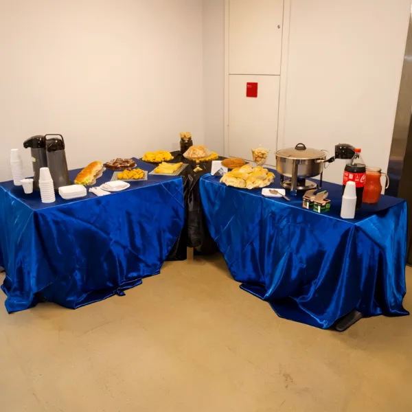 Duas mesas dispostas em formato de L, cobertas por toalhas azuis, servindo um buffet com garrafas térmicas, diversos pratos com salgados, sanduíches, um réchaud e bebidas, como refrigerante e suco